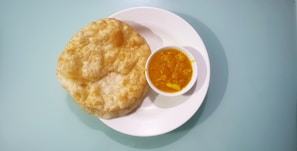 A white plate holds several puffed, golden-brown puris next to a small bowl of bright yellow curry with visible potato pieces.