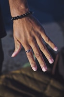 A close-up of a hand with a bracelet made of black beads and a ring featuring an infinity symbol. The skin tone is warm and the nails are neatly trimmed. The background is blurred, highlighting the hand as the focal point.