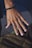 A close-up of a hand with a bracelet made of black beads and a ring featuring an infinity symbol. The skin tone is warm and the nails are neatly trimmed. The background is blurred, highlighting the hand as the focal point.