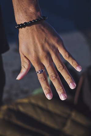 A close-up of a hand with a bracelet made of black beads and a ring featuring an infinity symbol. The skin tone is warm and the nails are neatly trimmed. The background is blurred, highlighting the hand as the focal point.