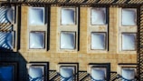 An aerial view of a tiled roof featuring multiple square-shaped skylights arranged in a grid pattern. The sunlight creates distinct shadows, highlighting the geometry and structure of the installation. The overall appearance suggests a utilitarian design focused on maximizing natural light entry into the space below.