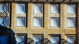 An aerial view of a tiled roof featuring multiple square-shaped skylights arranged in a grid pattern. The sunlight creates distinct shadows, highlighting the geometry and structure of the installation. The overall appearance suggests a utilitarian design focused on maximizing natural light entry into the space below.