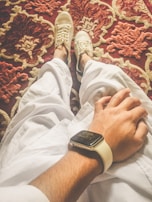 A person wearing white pants and white sneakers is sitting with one hand resting on the knee. The person is wearing a smartwatch on their wrist. The background features a richly patterned red and beige carpet.