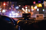 The interior of a car at night displays a blurred cityscape through the windshield, illuminated by various colorful lights. The dashboard is visible with a taximeter and an identification card prominently displayed on top. A person appears to be sitting in the front passenger seat, with a hand reaching toward the radio or dashboard.