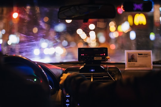 The interior of a car at night displays a blurred cityscape through the windshield, illuminated by various colorful lights. The dashboard is visible with a taximeter and an identification card prominently displayed on top. A person appears to be sitting in the front passenger seat, with a hand reaching toward the radio or dashboard.
