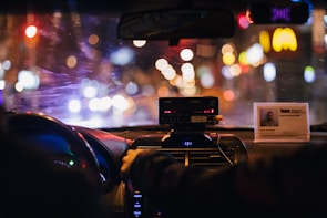 The interior of a car at night displays a blurred cityscape through the windshield, illuminated by various colorful lights. The dashboard is visible with a taximeter and an identification card prominently displayed on top. A person appears to be sitting in the front passenger seat, with a hand reaching toward the radio or dashboard.