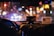 The interior of a car at night displays a blurred cityscape through the windshield, illuminated by various colorful lights. The dashboard is visible with a taximeter and an identification card prominently displayed on top. A person appears to be sitting in the front passenger seat, with a hand reaching toward the radio or dashboard.