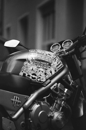 A close-up view of a motorcycle featuring a fuel tank covered with various stickers and decals. The image is in black and white, highlighting the intricate mechanical parts and gauges on the handlebar. The background is blurred, drawing focus to the bike&rsquo;s details.