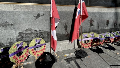 A memorial wall with etched images of military vehicles and soldiers serves as a backdrop to several large wreaths adorned with red poppies and purple ribbons. Two flags stand prominently next to the wreaths, casting shadows on the ground.