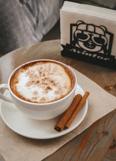 A cup of coffee with frothy milk and a sprinkle of cinnamon on top sits on a white saucer. Two cinnamon sticks are placed on the saucer beside the cup. A napkin holder with a cartoon bear wearing sunglasses is in the background on a wooden table.