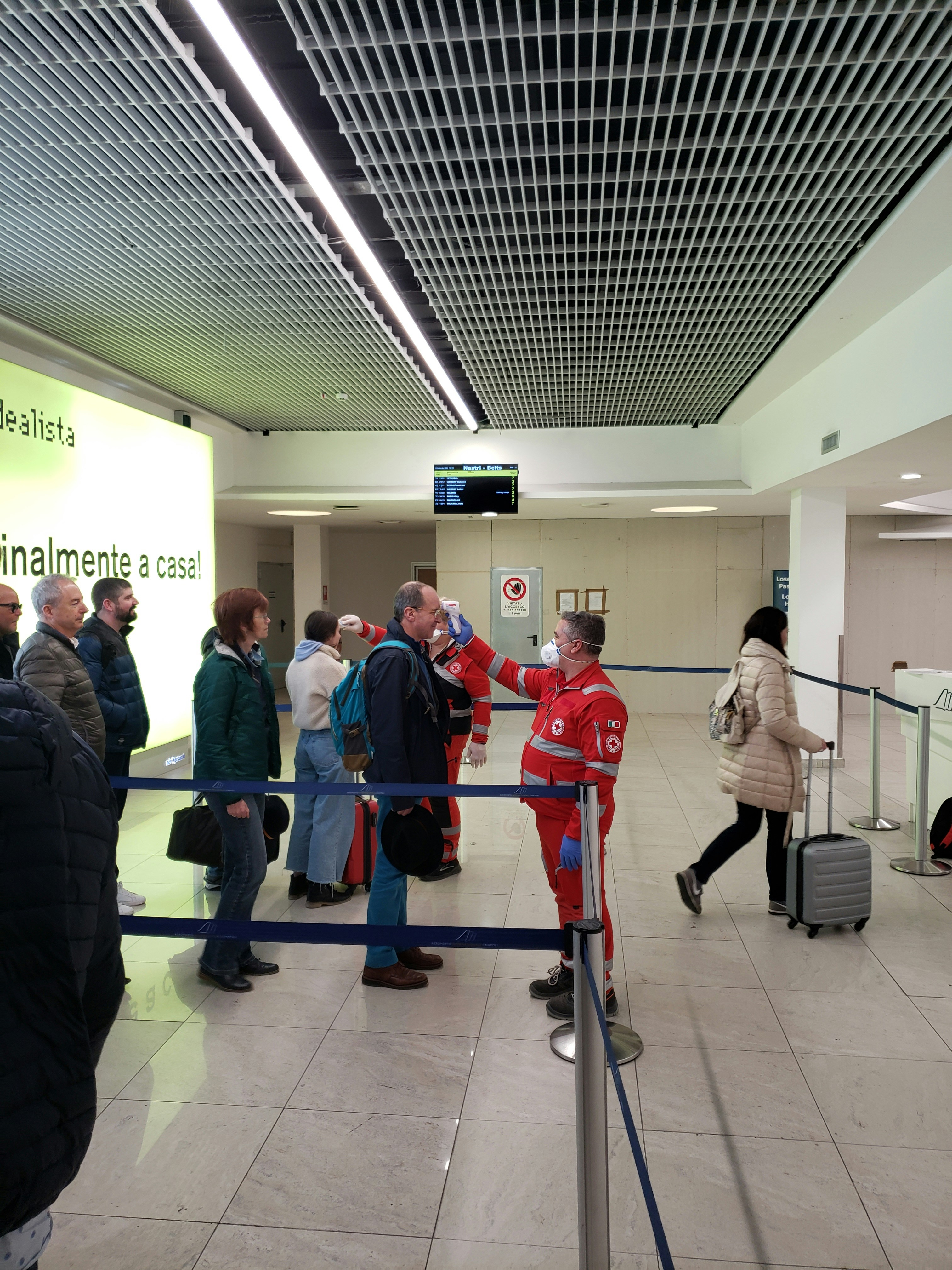 COVID-19 (coronavirus) temperature testing at Napoli Airport (Naples International Airport - NAP) by the Italian health authority (Ministero della Salute), wearing red. Thermometers are used in the head, workers wear n95 masks, passengers smile because they don't imagine what's going to happen in the coming months. Taken in February 2020, before all the lockdowns happened, when the pandemic was still beginning.