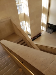 A modern, spacious interior with large, geometrically-designed stone stairs and walls. Sunlight streams through tall, narrow windows, casting shadows on the beige stone surfaces. A metal handrail accompanies the stairs, and in the lower area, several benches and an art display or installation with vertical metal rods can be seen.