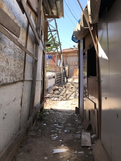 A narrow alleyway with makeshift walls constructed from wood and metal sheets. The corridor is cluttered with debris and scattered pieces of rubbish. Sunlight filters through gaps, casting shadows on the path. In the background, a series of similar constructed buildings can be observed along with some greenery.
