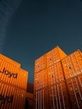 Stacks of bright orange shipping containers with the Hapag-Lloyd logo are arranged against a dark, overcast sky, creating a striking contrast. The containers are aligned neatly, showcasing international trade and logistics.