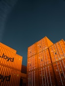 Stacks of bright orange shipping containers with the Hapag-Lloyd logo are arranged against a dark, overcast sky, creating a striking contrast. The containers are aligned neatly, showcasing international trade and logistics.