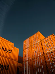 Stacks of bright orange shipping containers with the Hapag-Lloyd logo are arranged against a dark, overcast sky, creating a striking contrast. The containers are aligned neatly, showcasing international trade and logistics.