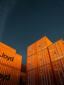 Stacks of bright orange shipping containers with the Hapag-Lloyd logo are arranged against a dark, overcast sky, creating a striking contrast. The containers are aligned neatly, showcasing international trade and logistics.