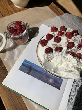 A round dessert topped with fresh raspberries and sunflower seeds sits on an open book. Nearby, a small glass jar is filled with more raspberries. Everything is placed on a piece of parchment paper on a wooden table, with natural sunlight casting shadows.