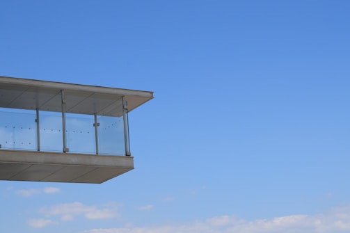 A modern structure with a glass balcony extends outwards against a clear blue sky. The architectural design features clean lines and transparency, creating a sense of openness.
