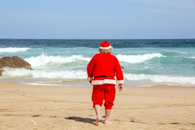 man in red dress shirt and brown shorts standing on beach during daytime santa claus teams background