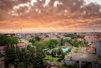 An urban landscape featuring a sprawling cityscape with diverse architectural styles. The foreground shows lush greenery, including tall trees and a park with pathways. Buildings of varying heights populate the middle and background under a dramatic sky painted with vibrant orange and yellow clouds, creating a picturesque sunset.