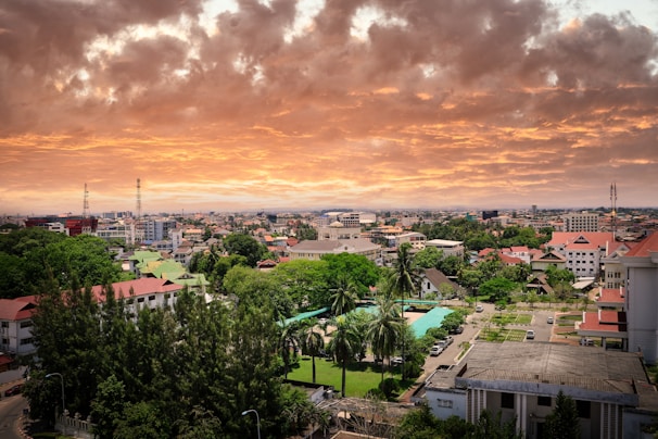 An urban landscape featuring a sprawling cityscape with diverse architectural styles. The foreground shows lush greenery, including tall trees and a park with pathways. Buildings of varying heights populate the middle and background under a dramatic sky painted with vibrant orange and yellow clouds, creating a picturesque sunset.