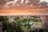 An urban landscape featuring a sprawling cityscape with diverse architectural styles. The foreground shows lush greenery, including tall trees and a park with pathways. Buildings of varying heights populate the middle and background under a dramatic sky painted with vibrant orange and yellow clouds, creating a picturesque sunset.