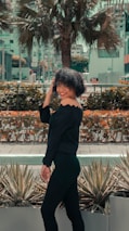 A woman with curly hair is smiling and standing in an outdoor setting. She is wearing an off-shoulder black outfit and appears to be posing near some agave plants. In the background, there are buildings and a large palm tree, surrounded by urban elements.
