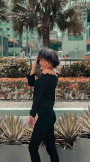 A woman with curly hair is smiling and standing in an outdoor setting. She is wearing an off-shoulder black outfit and appears to be posing near some agave plants. In the background, there are buildings and a large palm tree, surrounded by urban elements.