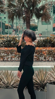A woman with curly hair is smiling and standing in an outdoor setting. She is wearing an off-shoulder black outfit and appears to be posing near some agave plants. In the background, there are buildings and a large palm tree, surrounded by urban elements.
