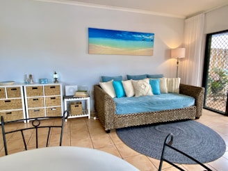 A cozy living room featuring a wicker daybed with blue and white cushions against a pastel-colored wall. Above the daybed hangs a beach-themed painting. To the left, a series of wicker drawers and decorative items add to the room’s decor. A soft rug lies on the tiled floor, and sheer white curtains partially cover a glass door, allowing natural light to brighten the space.