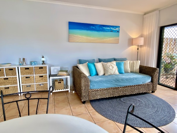 A cozy living room featuring a wicker daybed with blue and white cushions against a pastel-colored wall. Above the daybed hangs a beach-themed painting. To the left, a series of wicker drawers and decorative items add to the room’s decor. A soft rug lies on the tiled floor, and sheer white curtains partially cover a glass door, allowing natural light to brighten the space.