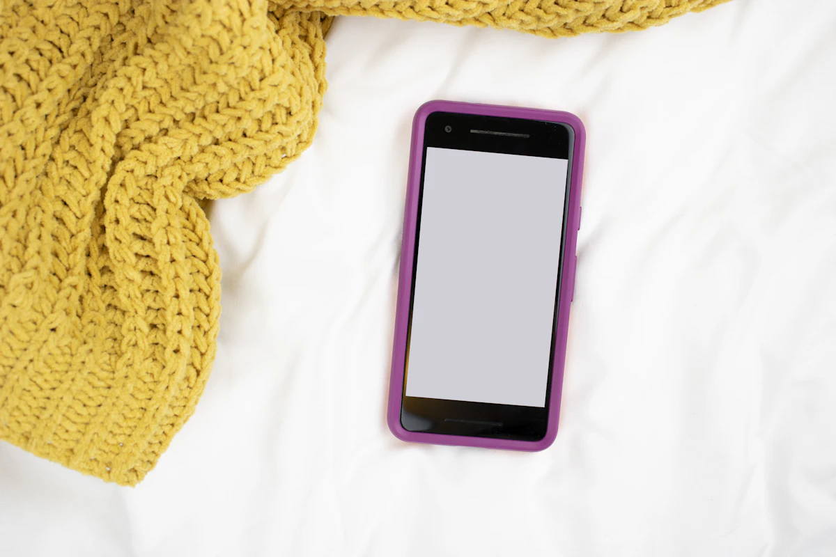 A phone with a purple case lying face up with a blank screen on white bedding, beside a mustard-yellow knitted blanket