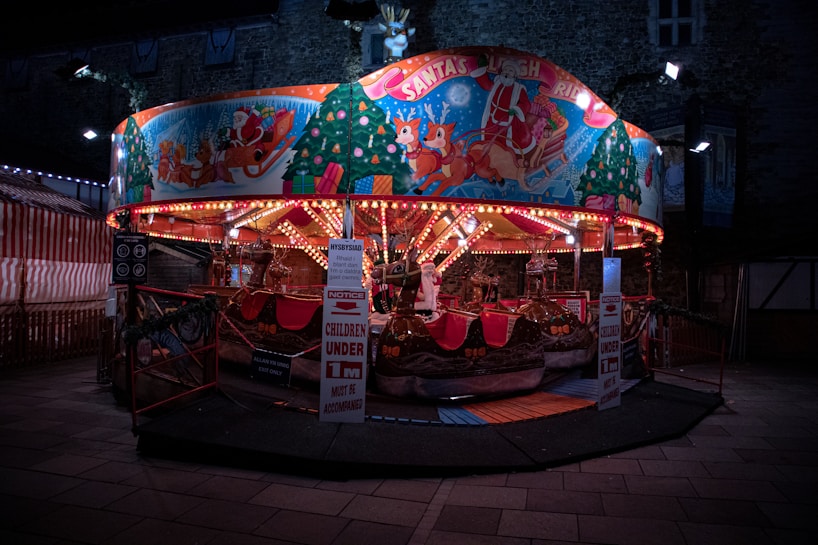 A festive amusement ride with colorful lights and a Christmas theme, featuring images of Santa Claus and reindeer on the canopy. The ride has several reindeer-shaped seats and is surrounded by bright, decorative bulbs. A sign is posted in front of the ride with safety notices for children.