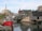 A small fishing village with wooden houses along a rocky shoreline. A red fishing boat labeled 'Harbour Mist' is docked in calm water. A Canadian flag is visible in the background, and the scene is serene with reflections in the water.