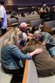 A group of people are gathered closely together in a church setting, engaged in prayer or a moment of reflection. Some are wearing masks, and they are seated on padded benches in what appears to be a worship area.