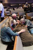 A group of people are gathered closely together in a church setting, engaged in prayer or a moment of reflection. Some are wearing masks, and they are seated on padded benches in what appears to be a worship area.