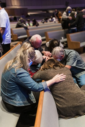 A group of people are gathered closely together in a church setting, engaged in prayer or a moment of reflection. Some are wearing masks, and they are seated on padded benches in what appears to be a worship area.