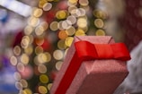 A glittery, pink gift box with a vibrant red bow is prominently displayed in the foreground. The background features a festive blur of circular, warm golden bokeh lights, suggesting a holiday or celebratory theme.