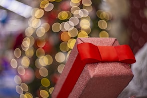 A glittery, pink gift box with a vibrant red bow is prominently displayed in the foreground. The background features a festive blur of circular, warm golden bokeh lights, suggesting a holiday or celebratory theme.