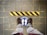 A pair of feet wearing brown shoes stand on a blue circular floor sign that reads 'STAND HERE', located on a tiled surface. In front of the feet is a yellow and black striped caution line adhered to the ground.