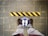 A pair of feet wearing brown shoes stand on a blue circular floor sign that reads 'STAND HERE', located on a tiled surface. In front of the feet is a yellow and black striped caution line adhered to the ground.