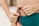Hands are tying a colorful braided string on a teal garment, with fingers adorned with delicate rings.