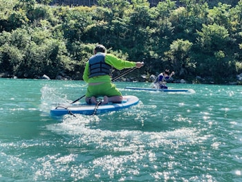 Two people are paddleboarding on a serene turquoise river. They are dressed in bright, sporty outfits and wearing life vests. The sun shines brightly, creating sparkling reflections on the water. A lush, green forest lines the riverbank in the background, adding to the natural beauty of the scene.