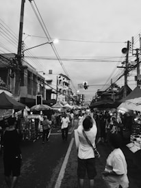A bustling street market lined with stalls on both sides, filled with people walking, shopping, and engaging with vendors. The street is busy with pedestrian activity, and power lines crisscross the sky above. A person in the foreground is taking a picture, while others are browsing merchandise or chatting.