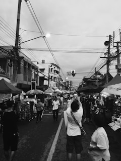 A bustling street market lined with stalls on both sides, filled with people walking, shopping, and engaging with vendors. The street is busy with pedestrian activity, and power lines crisscross the sky above. A person in the foreground is taking a picture, while others are browsing merchandise or chatting.