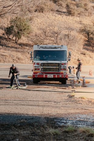 A red fire truck is parked on a road in a rural area with two firefighters preparing hoses. The surrounding landscape is dry with sparse trees, suggesting a dry or autumn season.