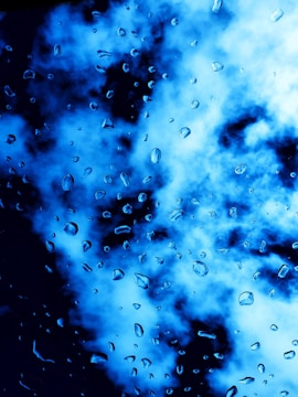 Water droplets are scattered across a surface, with a vibrant blue and black cloudy background. The droplets appear to be on glass, creating a dynamic and textured visual effect.