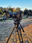 A professional camera mounted on a sturdy tripod is positioned outdoors in a natural setting. The background shows a grassy area with trees and a clear blue sky, indicating a sunny day.