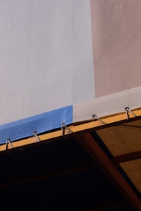 A close-up view of a building's facade with a combination of light blue and beige walls. The image features a part of a canopy or awning, which is predominantly blue with metal rings securing it to a yellow structure underneath, creating stark geometric patterns.